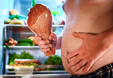 饥饿的胖子手里着条大火鸡腿,站打开的冰箱旁边健康的食物图片