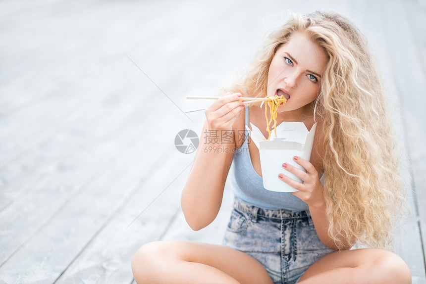 快乐的轻女人,留着长长的卷发,坐木地板上,着个饭盒,用筷子吃着中国外卖的条图片