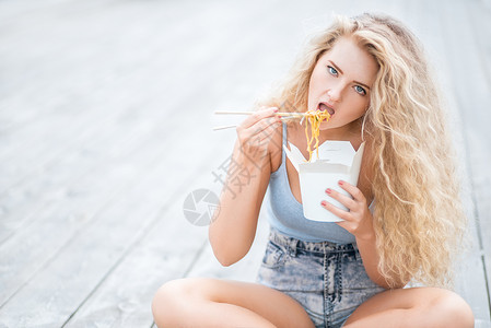 快乐的轻女人,留着长长的卷发,坐木地板上,着个饭盒,用筷子吃着中国外卖的条图片
