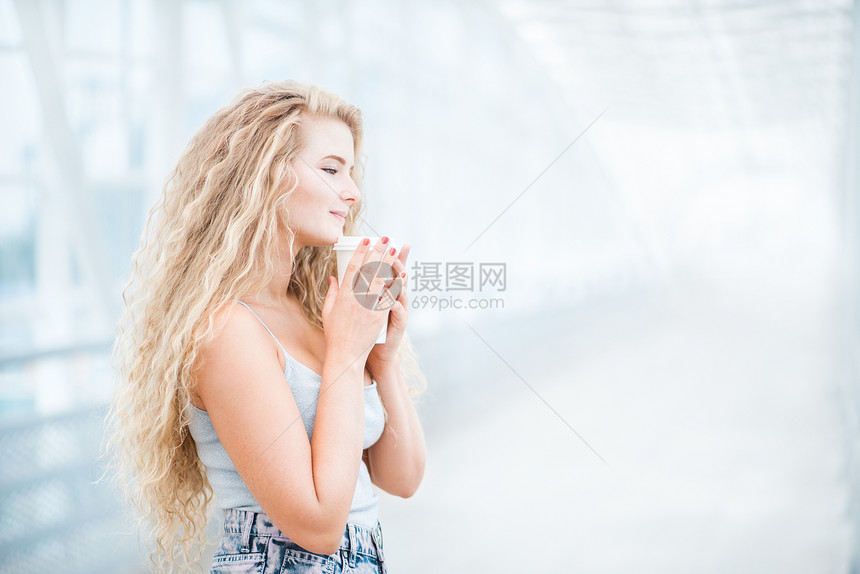 美丽的轻女人,留着长长的卷发,着个走的咖啡杯,站城市背景的桥上图片