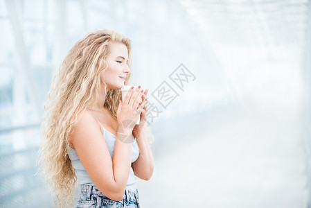 美丽的轻女人,留着长长的卷发,着个走的咖啡杯,站城市背景的桥上图片