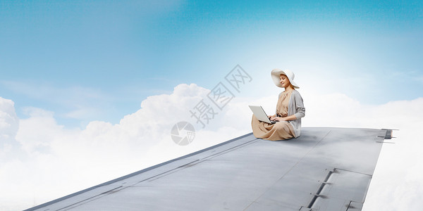 绝穿着连衣裙帽子的女人坐飞机机翼上,笔记本电脑上工作图片