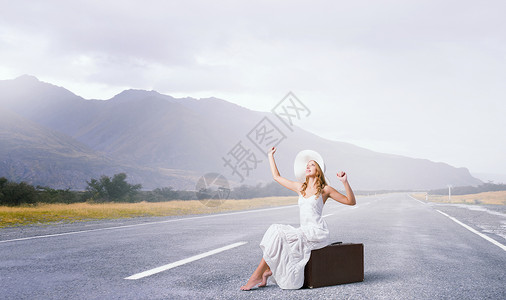 她旅行很轻穿着白色长裙帽子的女人坐沥青路上的行李上图片