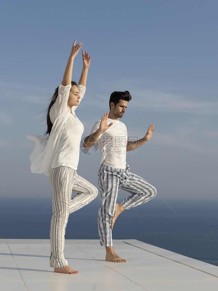 轻的夫妇日落时练瑜伽,现代的家庭梯田与海洋日落的背景图片