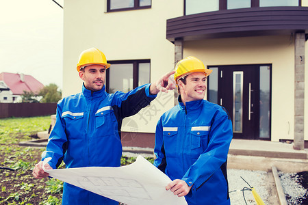 建筑,队合作人的两个微笑的建筑工人穿着硬帽子工作服,蓝图指着房子前的户外图片