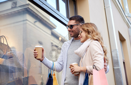 销售,消费主义人们的幸福的夫妇与购物袋咖啡纸杯看着城市的商店窗口图片