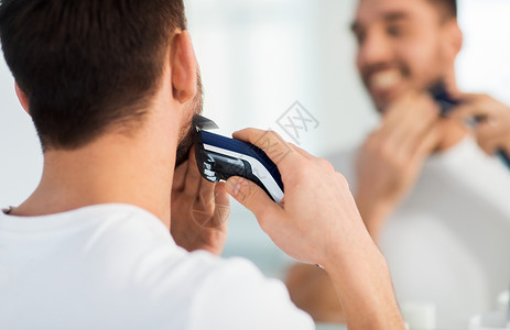 美容,剃须,美容人们的靠近轻人看镜子剃须胡须与修剪机电动剃须刀家里的浴室图片