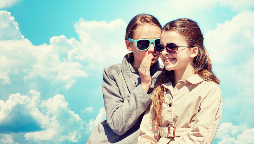 人,孩子友谊的快乐的小女孩戴着太阳镜,向朋友耳语她的秘密,蓝天云层背景上闲聊图片