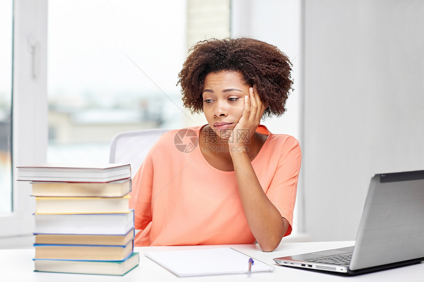 人技术教育理念无聊的非裔美国轻妇女坐桌子上,家里笔记本电脑书籍记事本图片