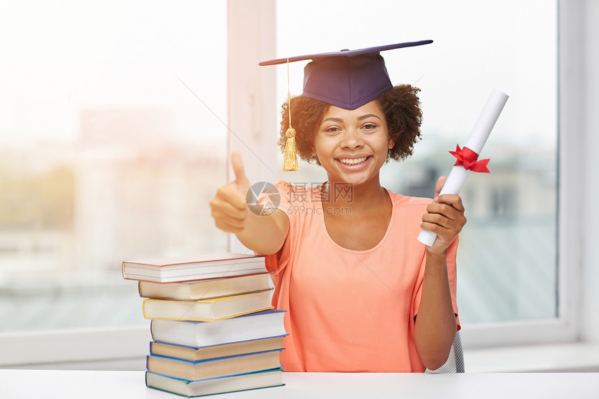 教育,学校,毕业,手势人的快乐的微笑非裔美国学生女孩戴着学士帽,书文凭坐桌子上,家里竖大拇指图片