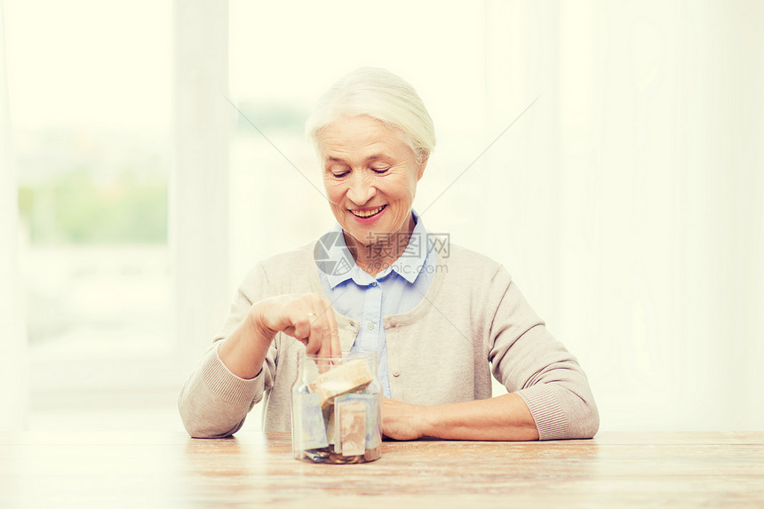 储蓄金钱金保险退休人们的观念微笑的老年女子家里把钞票放进璃瓶里图片