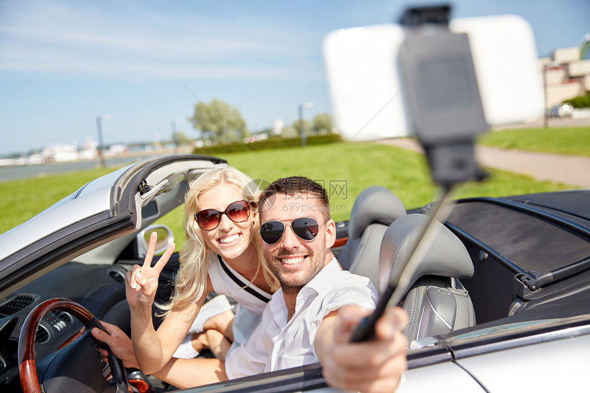 公路旅行,休闲,夫妇,技术人的快乐的男人女人驾驶敞篷车,并自拍杆上用智能手机拍照图片
