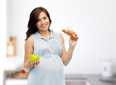 怀孕,健康饮食,垃圾食品人们的快乐的孕妇选择绿色苹果牛角包的家庭厨房背景图片