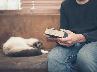 个轻人只睡猫坐沙发上,手里着本绿色的大书高清图片