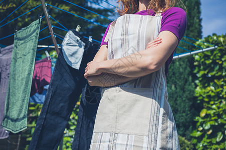 个穿着围裙的轻女人站她的洗衣房旁边,花园里的条晾衣绳上晾晒衣服图片