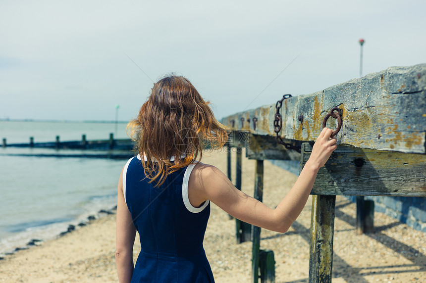 个穿着蓝色连衣裙的轻女人站海滩上,个阳光明媚的日子里,个古老的木制结构,挂着生锈的链子图片