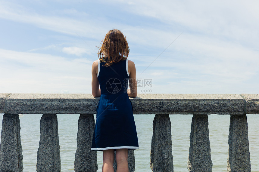 位穿着蓝色连衣裙的轻女子站长廊上的堵墙上,墙上挂着混凝土栏杆,个阳光明媚的日子里欣赏着海边图片