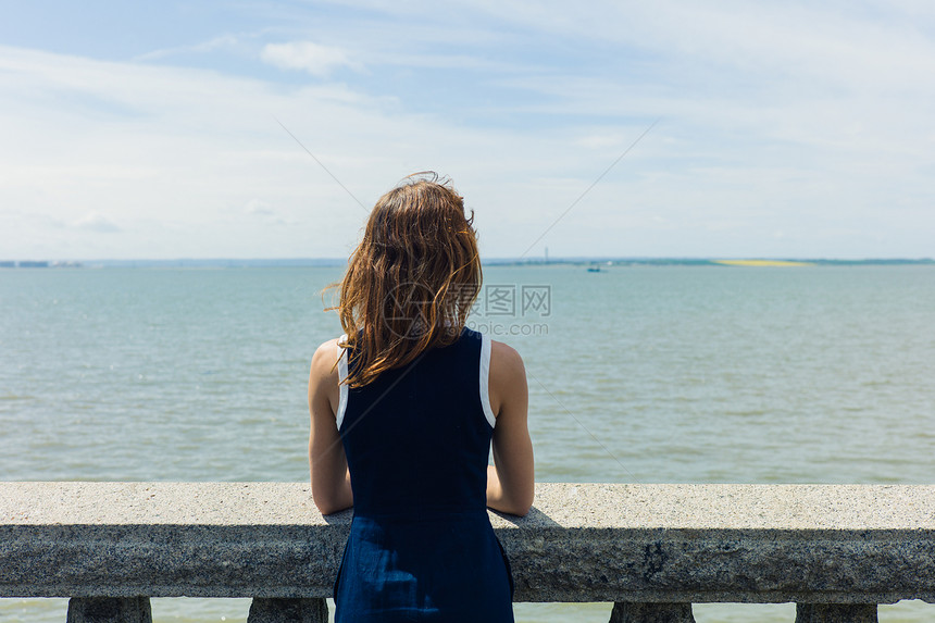 位穿着蓝色连衣裙的轻女子站长廊上的堵墙上,墙上挂着混凝土栏杆,个阳光明媚的日子里欣赏着海边图片