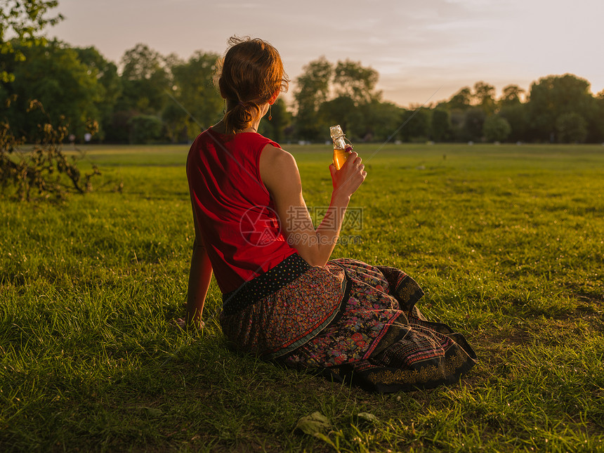 个轻的女人公园里坐草地上欣赏日落时,正瓶子里喝酒图片