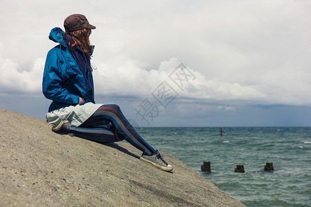 个穿着蓝色夹克的轻女人坐海边的防洪堤上图片
