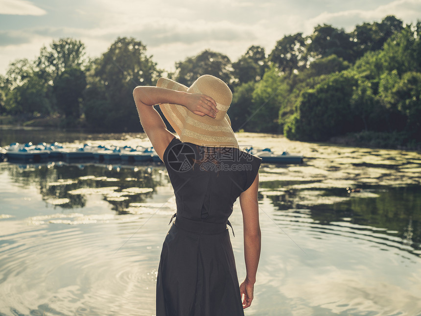位优雅的女人戴着顶帽子,日落时站公园的湖边,用老式的过滤镜头拍摄图片