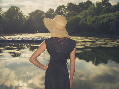 位优雅的女人戴着顶帽子,日落时站公园的湖边,用老式的过滤镜头拍摄图片