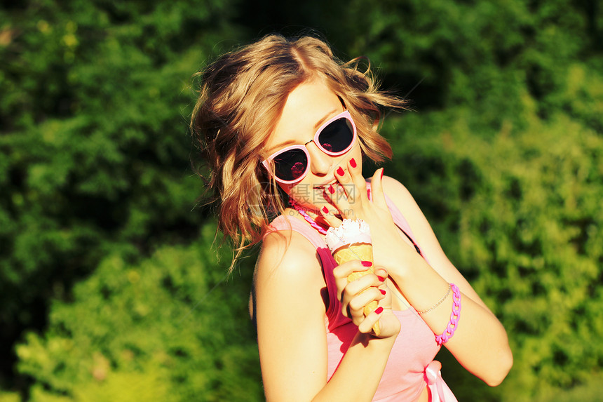 轻的金发时髦女孩夏天炎热的天气里吃着美味的冰淇淋,戴着太阳镜,心情愉快,镜头前微笑图片