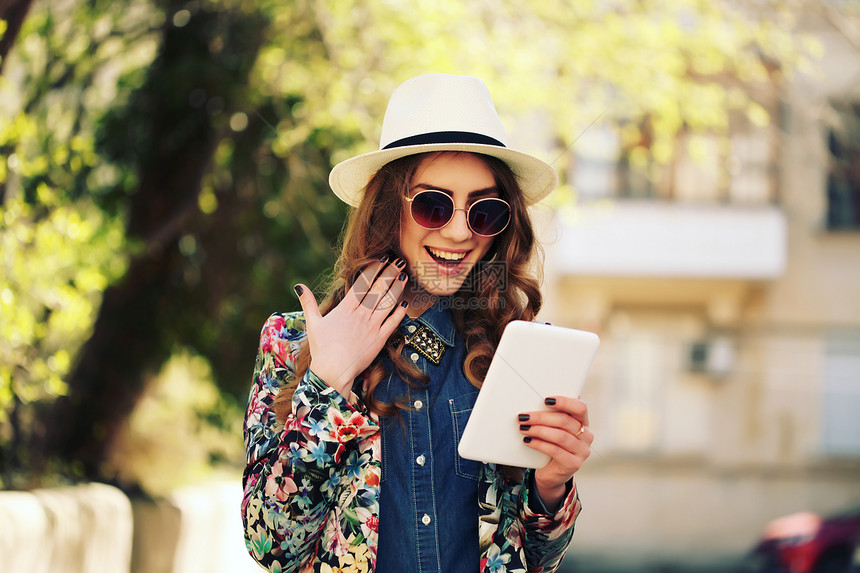 美丽微笑的时髦女人戴着老式太阳镜,牛仔裤衬衫,白色帽子夹克,用数码平板电脑给自己拍照自拍风格色调温暖的颜色户外图片