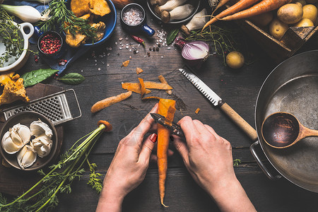 女女双手深色木制厨房桌子上剥胡萝卜,蔬菜烹饪原料勺子工具,俯视图背景图片