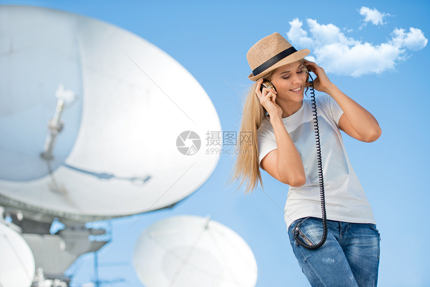 快乐的轻女子戴着帽子,老式音乐耳机中听音乐,接收卫星无线信号的卫星天线背景下跳舞图片