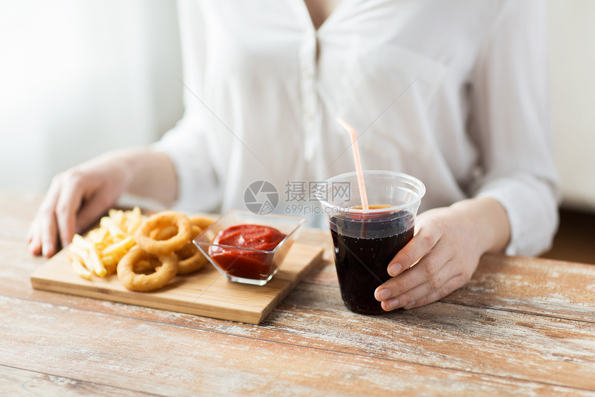 快餐,人健康的饮食观念密切妇女吃油炸鱿鱼戒指,炸薯条与番茄酱饮用可乐木桌上图片