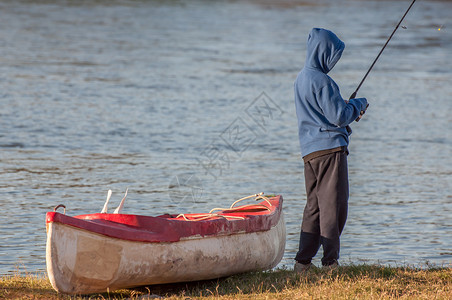 男孩,站河边的独木舟旁边,手里着鱼竿,忙着钓鱼高清图片