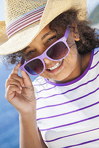个美丽而快乐的混血非裔美国女孩子戴着太阳镜牛仔帽蓝色热带海洋前度假图片