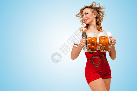 啤酒女郎轻的微笑感的瑞士女人穿着红色毛衣短裤,吊带的形式传统的Dirndl,着两个啤酒杯,边看着蓝色的背景背景
