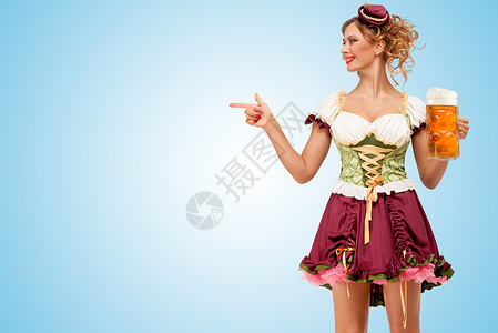 轻感的啤酒节女服务员穿着传统的巴伐利亚连衣裙,着个啤酒杯,指着蓝色的背景图片