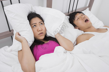 沮丧的女人用枕头盖住耳朵,而男人床上打鼾图片