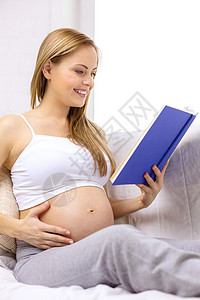 怀孕母微笑的孕妇坐沙发上,家看书图片