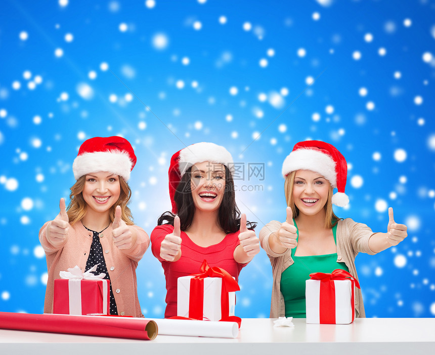 诞节,假日,手势,装饰人们的微笑的女人戴着诞老人助手帽子,装饰纸礼品盒,蓝色的雪背景上竖大拇指图片