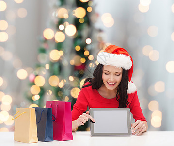 诞节,技术人的微笑的女人诞老人助手帽子与购物袋平板电脑电脑树灯背景图片