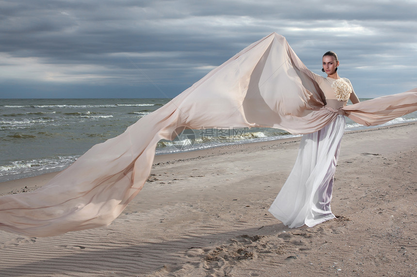 穿着米色长裙的女模特海滩上摆出动感十足的造型挥动布料飘动的长裙图片