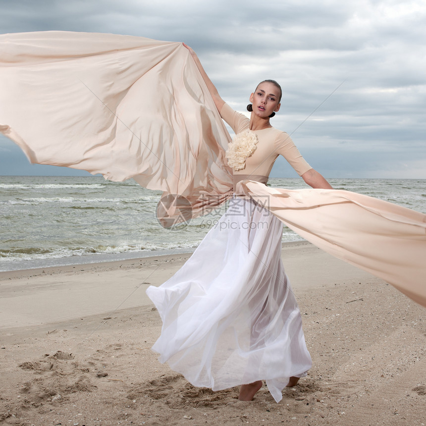 穿着米色长裙的女模特海滩上摆出动感十足的造型挥动布料飘动的长裙图片