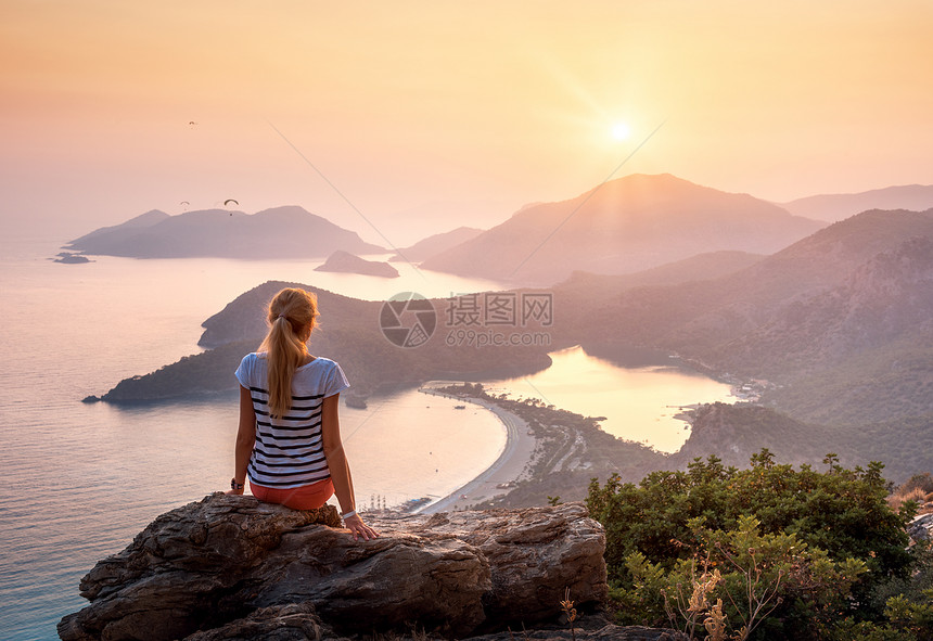 轻的女人坐岩石的顶部,看着海滩山脉,夏天五颜六色的日落风景与女孩,海洋,山脊橙色天空与太阳奥卢德尼斯,火鸡图片