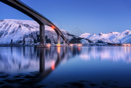 桥照明,雪覆盖着山,村庄蓝天,水丽的倒影夜间景观与桥梁,雪岩反射海上挪威洛福滕岛的冬天背景图片