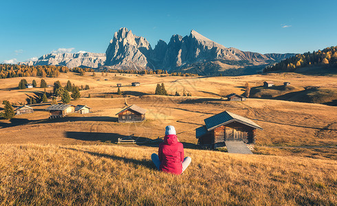 坐山上的女人秋天的日落时看着草地山脉阿尔佩迪西斯,白云石,意大利风景与女孩穿着红色夹克,田野与黄草,木屋岩石背景图片