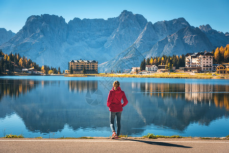秋天日出时,站米苏丽娜湖海岸的轻女子意大利白云石风景与女孩穿着红色夹克,倒影水,建筑物,蓝天意大利阿尔卑斯山背景