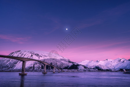 紫色夜桥美丽的桥抵雪山,紫色的天空,粉红色的云彩月亮夜间洛福滕岛,挪威冬天的风景道路,蓝色的大海,雪覆盖的岩石黄昏欧洲背景