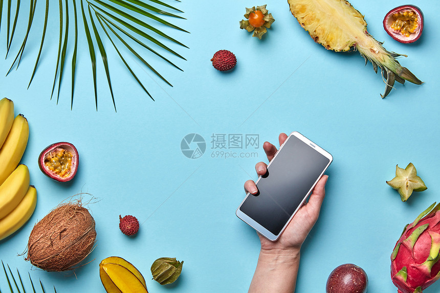 棕榈叶,椰子,荔枝,菠萝套热带水果女孩的手着蓝色背景的手机,个的副本现代网上购物的平躺各种热图片