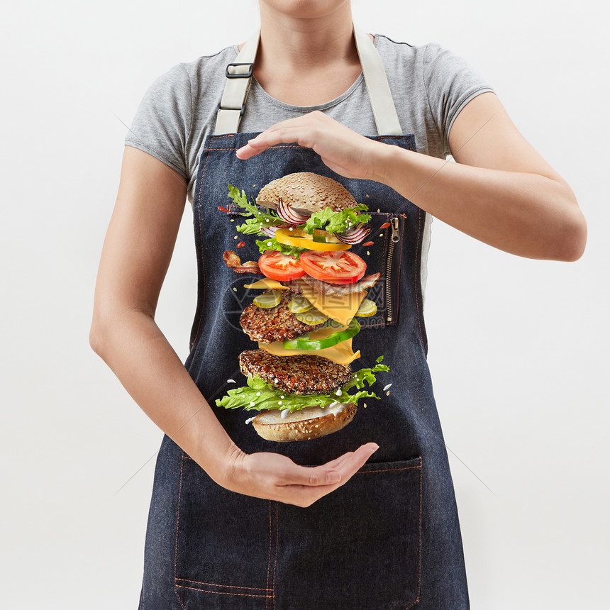 女人厨师穿着牛仔围裙,手里着白色背景上新鲜天然食材的美味自制汉堡文字的地方大自制的新鲜汉堡女孩的手中,飞行的成分图片