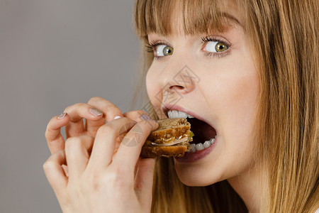 轻的女人吃三明治,用张开的嘴咬人食物,卡路里,节食的工作室拍摄灰色背景,侧写视图女人吃三明治,咬人背景图片