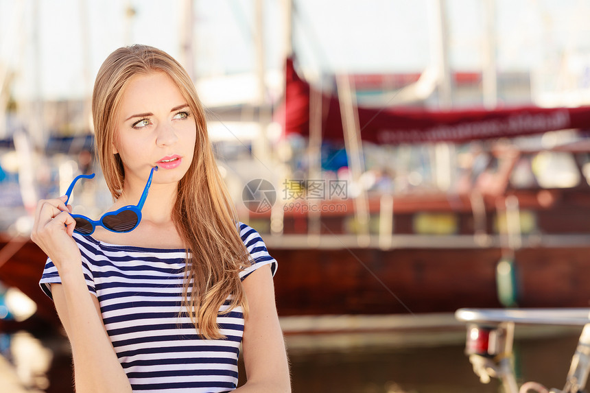 旅游人的时尚金发女孩带着蓝色心形太阳镜码头抗游艇港口图片